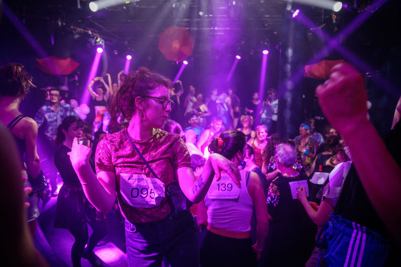 Salle remplie de danseurs et danseuses. Au premier plan une danseuse en action portant un dossart.
